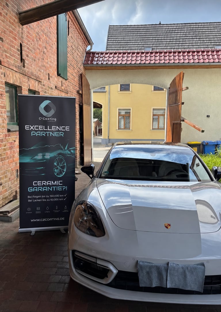 White Porsche sports car parked in front of a ceramic coating service sign at an auto detailing facility with brick buildings in background